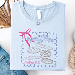 a light blue t-shirt with a graphic design featuring bread and fish, along with a pink ribbon. The t-shirt is laid out on a white background, accompanied by a pair of jeans, a pair of white sneakers, and a beige sweater.