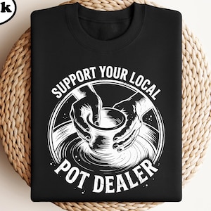 May include: Black crew neck t-shirt with a white graphic. The graphic features hands shaping a pot on a pottery wheel, encircled by the text "SUPPORT YOUR LOCAL POT DEALER". The shirt is displayed on a woven, circular surface.