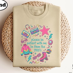 May include: Sand-colored sweatshirt with a retro-themed graphic. The design includes a pink star, cassette tape, boombox, roller skate, and the text "please be patient with me I'm from the 1900's".