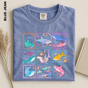May include: A blue jean colored t-shirt featuring a graphic print of various sharks in vibrant colors including pink, yellow, teal, and purple. The sharks are arranged in rectangular and heart-shaped frames with small star and wave accents.