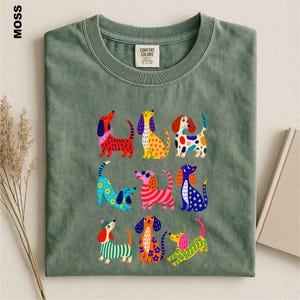 May include: A moss green crew neck t-shirt featuring a graphic print of nine stylized dogs in various colors and patterns. The shirt has a Comfort Colors brand label at the inner neckline. The dogs are depicted with spots, stripes, and floral designs.