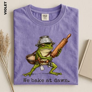 Op de afbeelding: Een violet t-shirt met een afbeelding van een groene kikker die een vergiet als hoed draagt en een houten deegroller en een kom met deeg vasthoudt. De kikker draagt een riem met keukengerei. De tekst onderaan luidt, We bake at dawn.