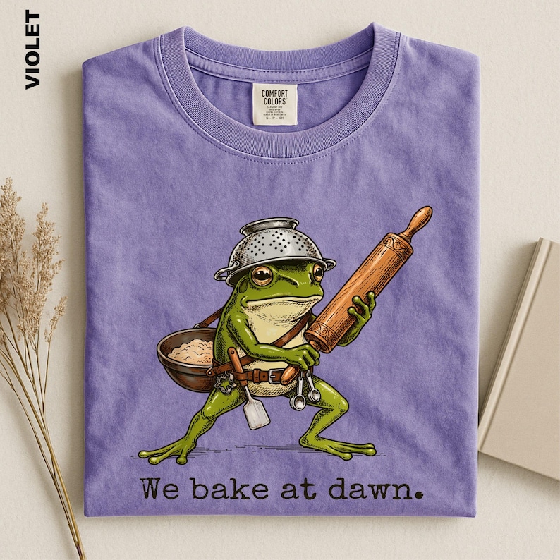 Op de afbeelding: Een violet t-shirt met een afbeelding van een groene kikker die een vergiet als hoed draagt en een houten deegroller en een kom met deeg vasthoudt. De kikker draagt een riem met keukengerei. De tekst onderaan luidt, We bake at dawn.