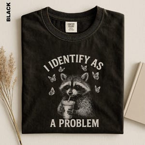 May include: A black crew neck t-shirt featuring a white graphic illustration of a raccoon holding a drink cup with a straw. The text "I IDENTIFY AS A PROBLEM" is printed in white above and below the raccoon, surrounded by small butterfly icons.