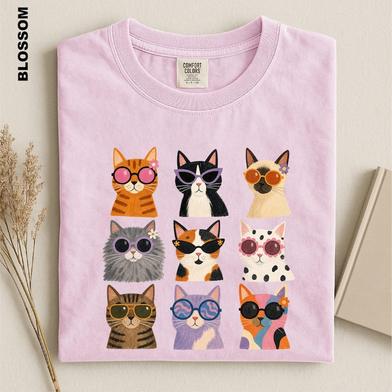 May include: A light purple t-shirt featuring a graphic print of nine cats wearing various colorful sunglasses and small flower accessories. The shirt has a crew neckline and a Comfort Colors brand label on the inside of the collar.