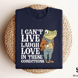 May include: A navy blue t-shirt with a white graphic of a frog holding a cup of coffee. The text on the shirt reads "I can't live laugh love in these conditions."