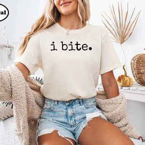 A woman wearing a white t-shirt with the text "i bite" printed on it, paired with denim shorts, sits in a room with a woven basket and a plant.