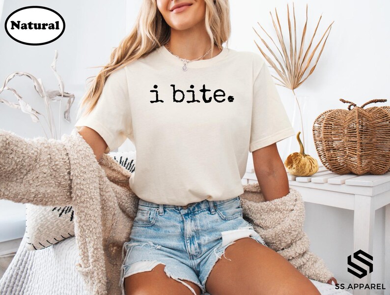 A woman wearing a white t-shirt with the text "i bite" printed on it, paired with denim shorts, sits in a room with a woven basket and a plant.