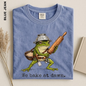 Op de afbeelding: Een blauwkleurig t-shirt met een afbeelding van een groene kikker die een metalen vergiet als hoed draagt. De kikker houdt een houten deegroller vast en draagt een bruine kom met een spatel en maatlepels. De tekst op het shirt is: We bake at dawn.