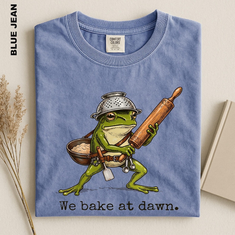 Op de afbeelding: Een blauwkleurig t-shirt met een afbeelding van een groene kikker die een metalen vergiet als hoed draagt. De kikker houdt een houten deegroller vast en draagt een bruine kom met een spatel en maatlepels. De tekst op het shirt is: We bake at dawn.