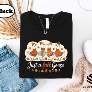 a black t-shirt with a graphic design of four geese wearing orange and yellow scarves, along with the text "Just a fall Goose" printed on it. The t-shirt is laid out on a white background, accompanied by a pair of blue jeans, a pair of white sneakers, and some leaves.