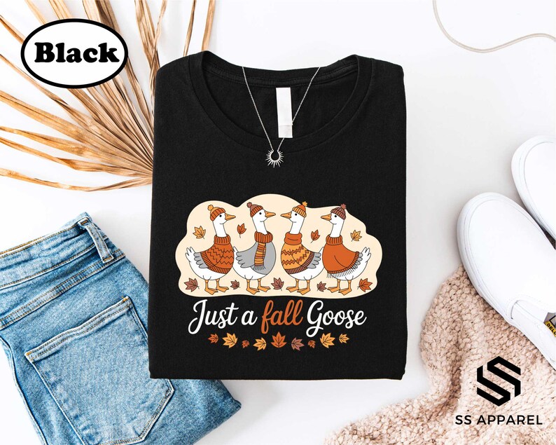 a black t-shirt with a graphic design of four geese wearing orange and yellow scarves, along with the text "Just a fall Goose" printed on it. The t-shirt is laid out on a white background, accompanied by a pair of blue jeans, a pair of white sneakers, and some leaves.