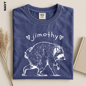May include: A navy blue t-shirt featuring a white graphic of a raccoon walking. Above the raccoon, the text "jimothy" is printed in a white, handwritten-style font, flanked by two small white heart outlines. The shirt has a crew neckline.