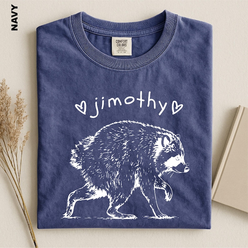May include: A navy blue t-shirt featuring a white graphic of a raccoon walking. Above the raccoon, the text "jimothy" is printed in a white, handwritten-style font, flanked by two small white heart outlines. The shirt has a crew neckline.