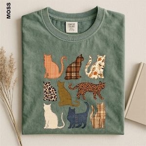 May include: A moss green crew neck t-shirt featuring a graphic print of nine cats in various patterns, including plaid, leopard print, and solid colors. The shirt has a Comfort Colors brand label at the inner neckline and a short-sleeved design.