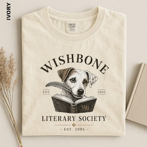 a beige t-shirt with a graphic design depicting a dog holding a book, along with text that reads "Wishbone Literary Society" and "EST. 1995 - EST. 1995".