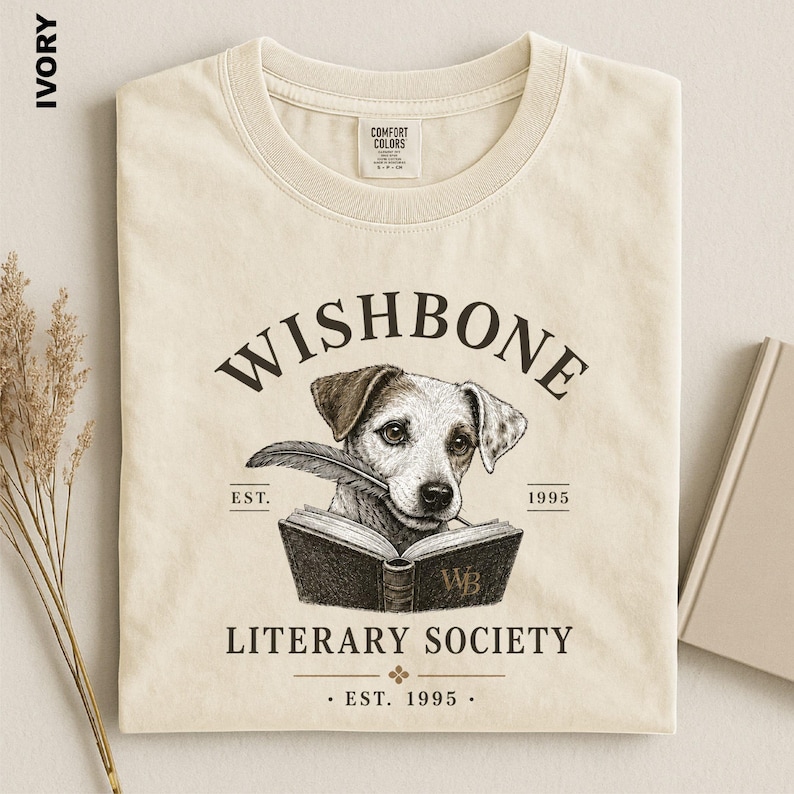 a beige t-shirt with a graphic design depicting a dog holding a book, along with text that reads "Wishbone Literary Society" and "EST. 1995 - EST. 1995".