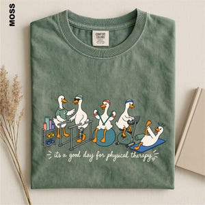 May include: A moss green crew neck t-shirt featuring a graphic of five white ducks performing physical therapy exercises. The text below reads, "it's a good day for physical therapy." The shirt has a Comfort Colors brand label at the inner neckline.