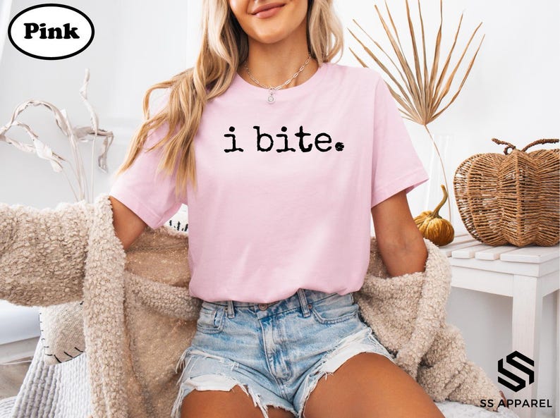 A woman wearing a pink t-shirt with the text "i bite" printed on it, sitting in a chair with a beige sweater draped over her.
