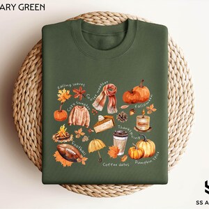 May include: A green t-shirt with a fall-themed design featuring illustrations of pumpkins, leaves, a cozy sweater, a coffee cup, and the text "Falling leaves", "Cozy sweater", "Cooler weather", "Halloween", "Thanksgiving", "Pumpkin spice", and "Coffee dates".