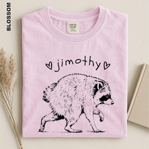 May include: A light pink t-shirt featuring a black line-art illustration of a raccoon. Above the raccoon, the text reads "jimothy" in a handwritten font, flanked by two small heart symbols. The shirt has a Comfort Colors brand neck label.