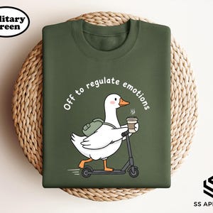 May include: Military green sweatshirt featuring a cartoon goose riding a scooter, carrying a backpack and a coffee cup. The text "Off to regulate emotions" is arched above the goose. The sweatshirt has a crew neck and long sleeves.