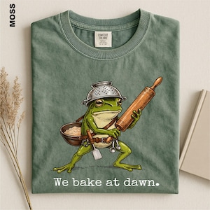Op de afbeelding: Een mosgroen t-shirt met een afbeelding van een groene kikker die een vergiet als hoed draagt, een houten deegroller vasthoudt en een kom en keukengerei bij zich heeft. De tekst "We bake at dawn." staat in het wit onder de illustratie.