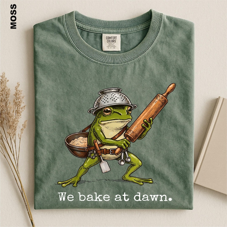 Op de afbeelding: Een mosgroen t-shirt met een afbeelding van een groene kikker die een vergiet als hoed draagt, een houten deegroller vasthoudt en een kom en keukengerei bij zich heeft. De tekst "We bake at dawn." staat in het wit onder de illustratie.