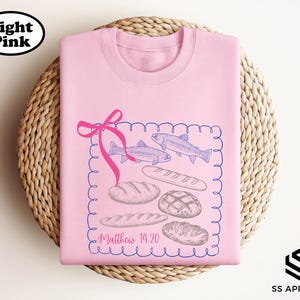 A pink t-shirt with a graphic design featuring fish, bread, and a ribbon, along with the text "Matthew 14:20" printed on it.