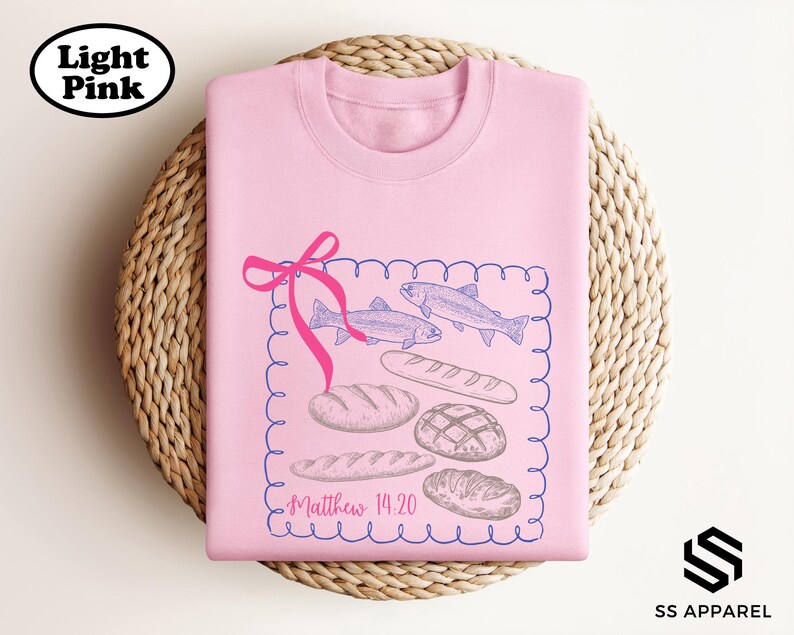 A pink t-shirt with a graphic design featuring fish, bread, and a ribbon, along with the text "Matthew 14:20" printed on it.