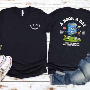a black t-shirt with a graphic design and text, along with a pair of jeans and sneakers, all laid out on a wooden surface.