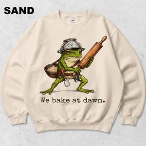 Op de afbeelding: Een zandkleurige sweater met ronde hals en een afbeelding van een groene kikker die een vergiet als helm draagt, een houten deegroller en een kom vasthoudt, met de tekst "We bake at dawn." in zwarte letters onder de illustratie.