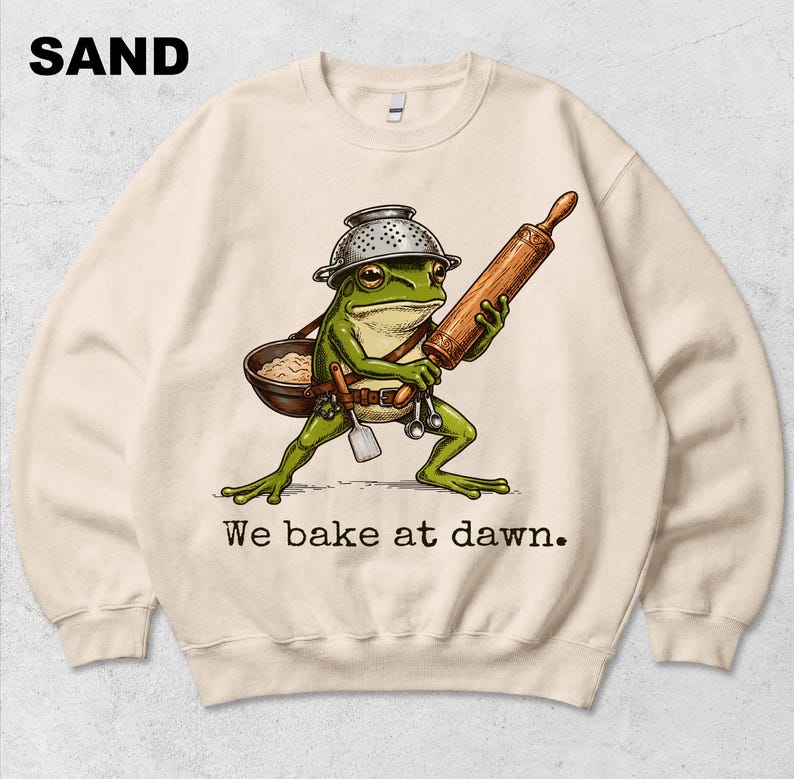 Op de afbeelding: Een zandkleurige sweater met ronde hals en een afbeelding van een groene kikker die een vergiet als helm draagt, een houten deegroller en een kom vasthoudt, met de tekst "We bake at dawn." in zwarte letters onder de illustratie.