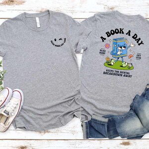 a gray t-shirt with a cartoon character and text printed on it, along with a pair of jeans and sneakers, all laid out on a wooden surface.
