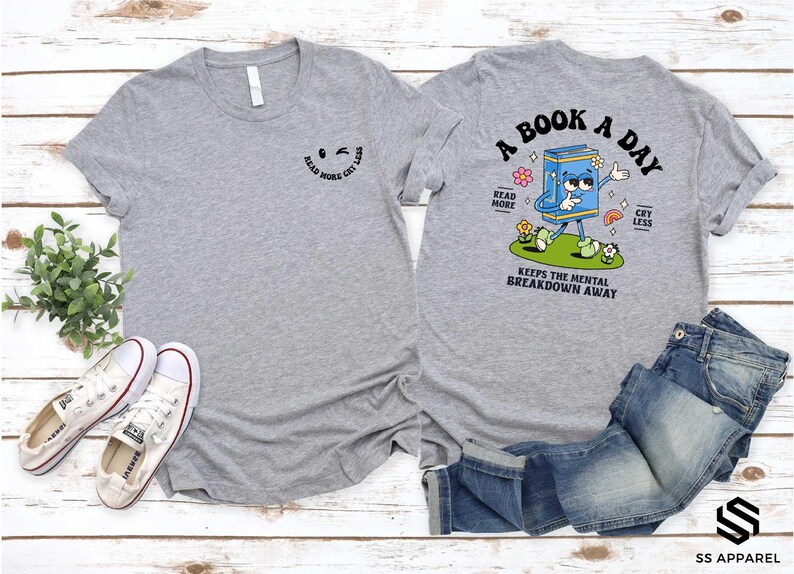 a gray t-shirt with a cartoon character and text printed on it, along with a pair of jeans and sneakers, all laid out on a wooden surface.