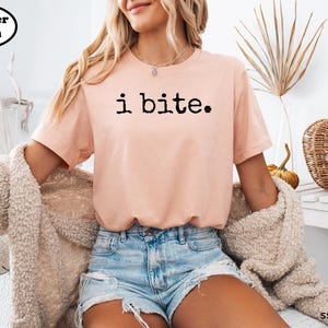 A woman wearing a pink t-shirt with the text "i bite" is sitting in a chair, with a beige sweater draped over her.