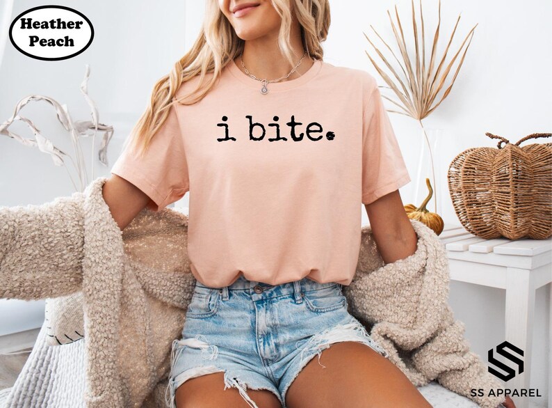 A woman wearing a pink t-shirt with the text "i bite" is sitting in a chair, with a beige sweater draped over her.