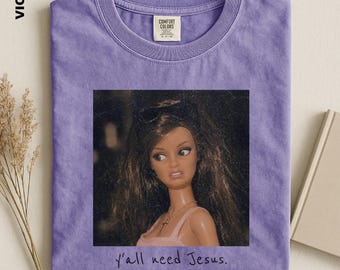 May include: A violet crew neck t-shirt featuring a graphic print of a doll with long brown hair wearing sunglasses and a necklace. The text "y'all need Jesus." is printed below the doll image. The shirt has a Comfort Colors brand label.