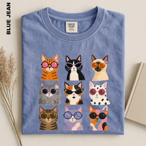 May include: A blue jean colored t-shirt featuring a graphic print of nine cats arranged in a grid. Each cat wears colorful sunglasses and some have small flowers on their heads. The shirt has a crew neckline and a Comfort Colors brand label.
