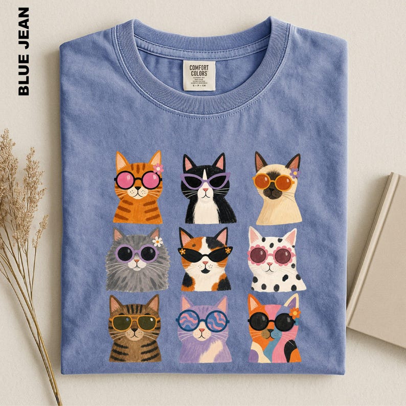May include: A blue jean colored t-shirt featuring a graphic print of nine cats arranged in a grid. Each cat wears colorful sunglasses and some have small flowers on their heads. The shirt has a crew neckline and a Comfort Colors brand label.