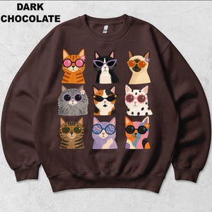 May include: A dark chocolate colored crewneck sweatshirt featuring a graphic print of nine cats arranged in a grid. Each cat wears colorful sunglasses and some have small flowers near their ears. The text DARK CHOCOLATE is printed in black.