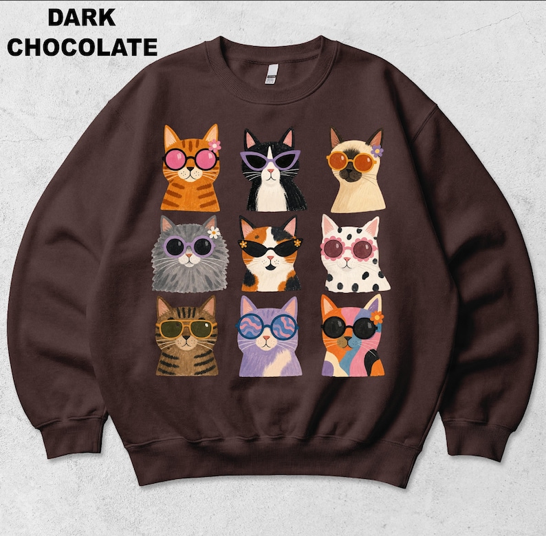 May include: A dark chocolate colored crewneck sweatshirt featuring a graphic print of nine cats arranged in a grid. Each cat wears colorful sunglasses and some have small flowers near their ears. The text DARK CHOCOLATE is printed in black.
