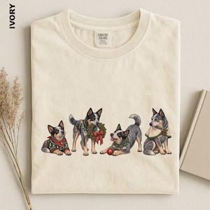 May include: An ivory crew neck t-shirt featuring a graphic of four Blue Heeler dogs. One dog wears a green jumper, one holds a holiday wreath, one plays with a red ornament while wearing a collar, and one wears a cape. The shirt has a Comfort Colors label.