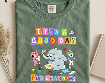 May include: A moss green t-shirt featuring a colorful graphic with the text IT'S A GOOD DAY FOR LEARNING. The design includes a cartoon elephant drawing, a pig holding books, a rabbit reading, and a bird, surrounded by various school supplies.