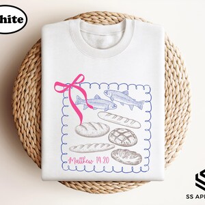 A white t-shirt with a graphic design featuring illustrations of bread and fish, along with the text "Matthew 14:20" printed on it.