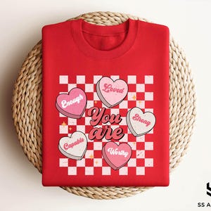 May include: Red t-shirt with a checkered pattern and pink hearts that say "Loved", "Enough", "Strong", "Capable", and "Worthy". The text "You are" is written in a retro font.