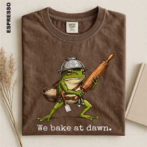 Op de afbeelding: Een espresso-kleurig t-shirt met een afbeelding van een groene kikker die een vergiet als hoed draagt, een houten deegroller vasthoudt en een kom met deeg draagt. De tekst "We bake at dawn." staat in het wit onder de kikkerillustratie.