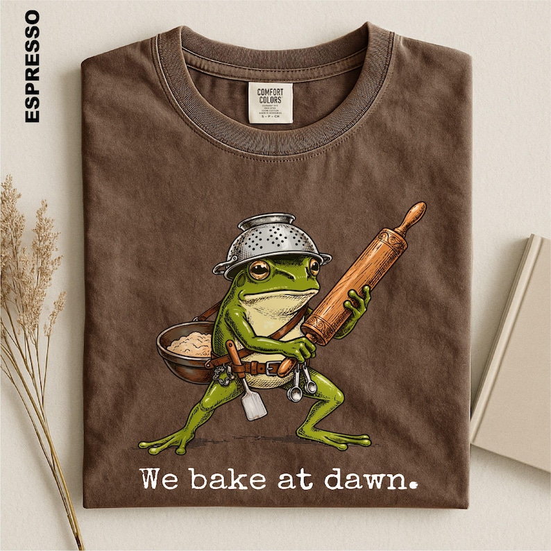Op de afbeelding: Een espresso-kleurig t-shirt met een afbeelding van een groene kikker die een vergiet als hoed draagt, een houten deegroller vasthoudt en een kom met deeg draagt. De tekst "We bake at dawn." staat in het wit onder de kikkerillustratie.