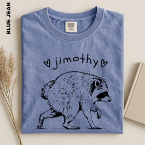 May include: A blue jean colored t-shirt featuring a black line art illustration of a raccoon walking. Above the raccoon, the text reads "jimothy" in a lowercase handwritten font, flanked by two small black heart symbols on either side.