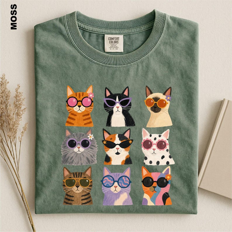 May include: A moss green crew neck t-shirt featuring a graphic print of nine cats arranged in a grid. Each cat wears colorful sunglasses and some have small flowers on their heads. The shirt has a Comfort Colors brand label at the inner neckline.